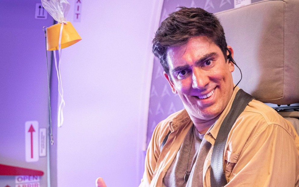 Marcelo Adnet em um avião; humorista sorri de lado preso a um cinto de segurança de avião