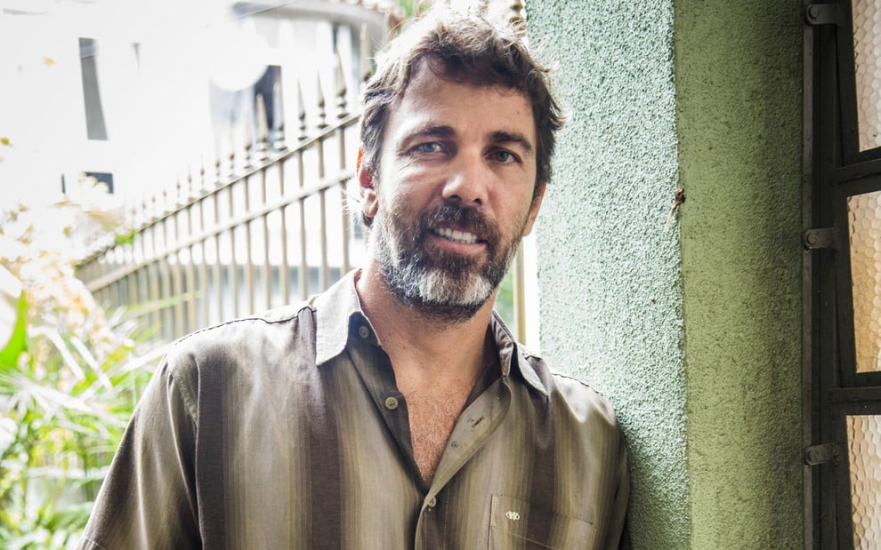 Marcelo Faria posa com camisa listrada bege e marrom caracterizado como seu personagem da novela Bom Sucesso