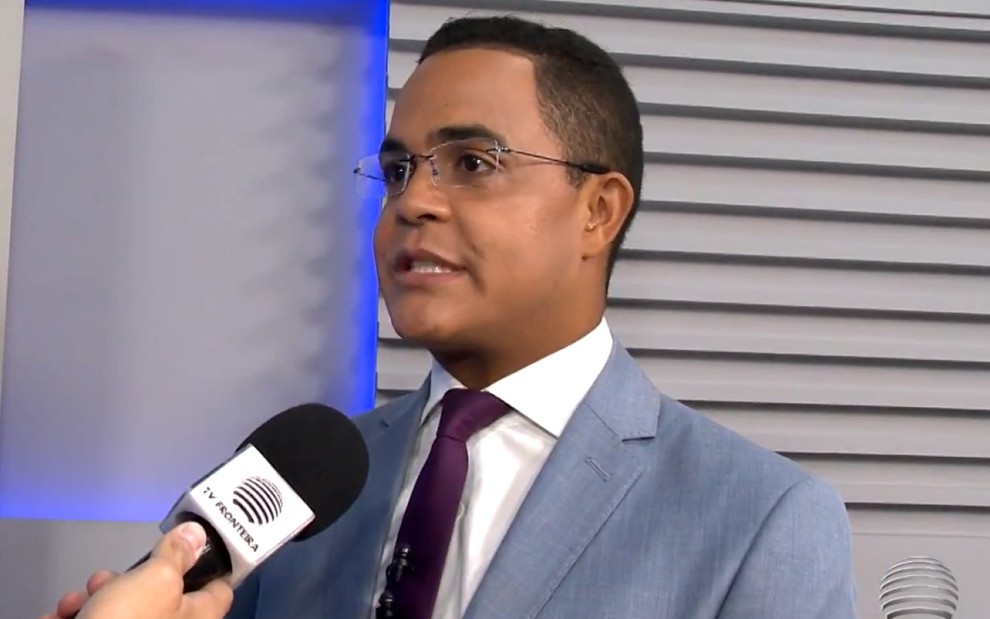 Marcelo Pereira de terno cinza claro, camisa branca e gravata bordô falando ao microfone em um estúdio de televisão