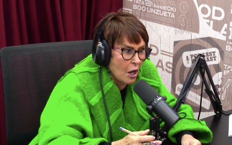 A espiritualista Márcia Sensitiva com um casaco verde com um fone de ouvido em frente a um microfone no estúdio do PodDelas