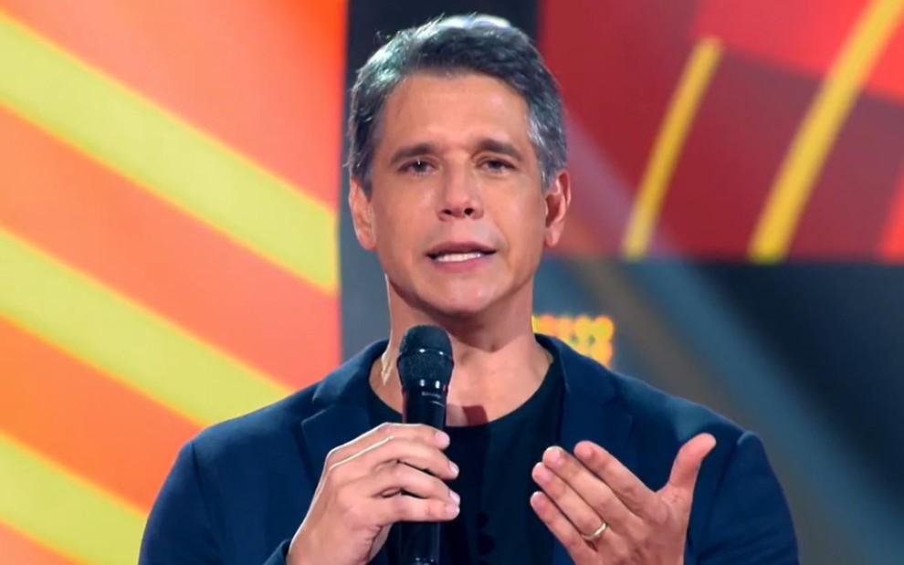 Marcio Garcia no comando da sétima temporada do The Voice Kids; ele segura o microfone e fala para a câmera