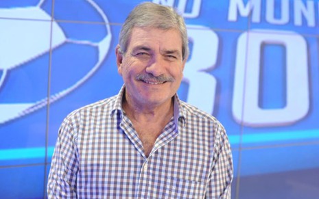 Márcio Guedes usa uma camia social listrada e sorri para a câmera; ao fundo, é possível ver o cenário do programa No Mundo da Bola