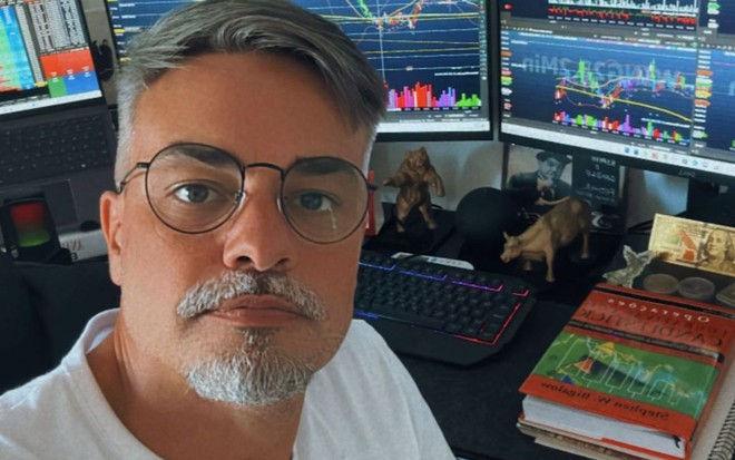 O ator Márcio Kieling em frente a monitores, com camiseta branca, óculos e expressão séria