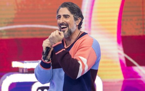 Marcos Mion usa uma camisa quadriculada, sorri e fala com o público do Caldeirão em sua estreia na Globo