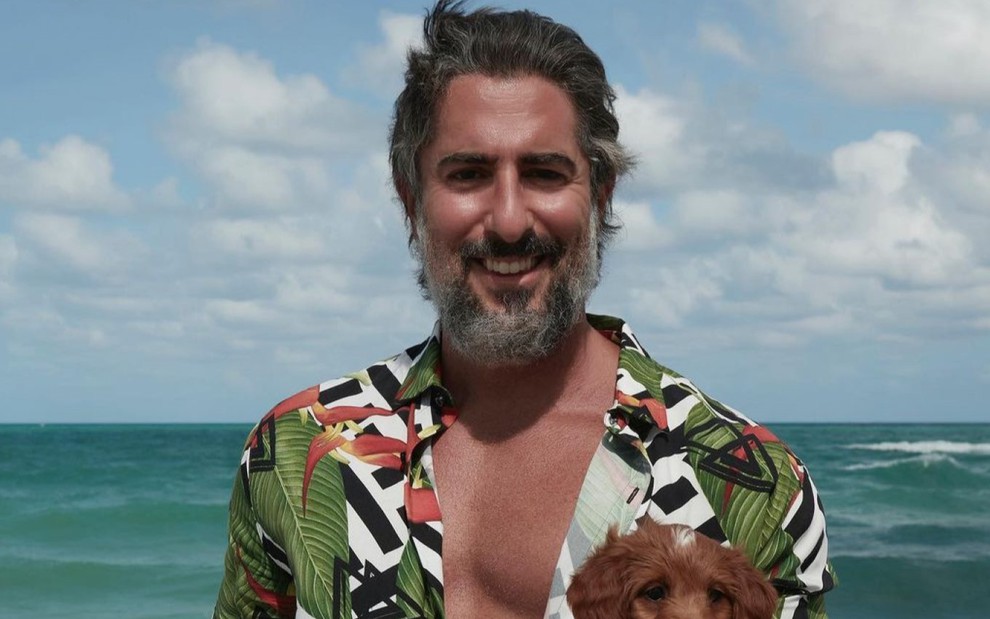 Marcos Mion com uma camisa florida, na praia, segurando um cachorrinho