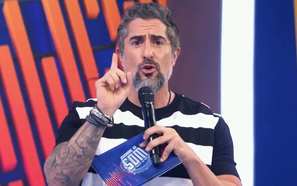Marcos Mion faz sinal de 1 na mão enquanto segura o microfone para apresentar o Caldeirão; ele está de camiseta