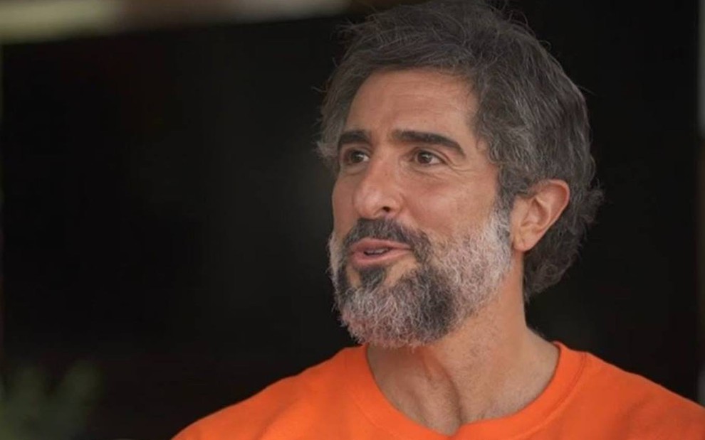 Marcos Mion usa uma camisa laranja, sorri e recebe a equipe do Fantástico em sua casa, no interior de São Paulo, onde foi apresentado para o público da Globo