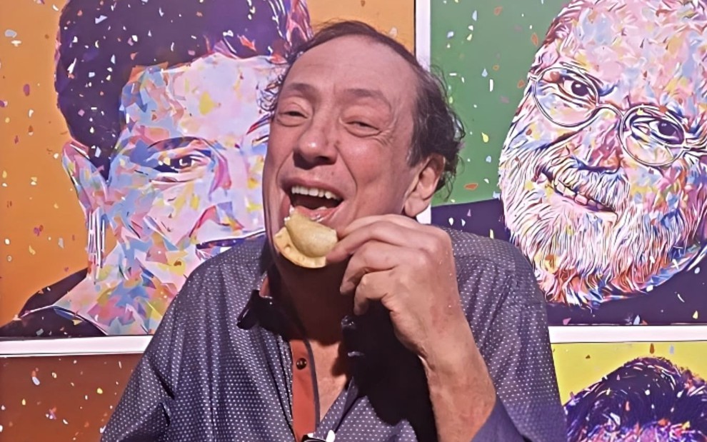 Marcos Oliveira posa sorrindo e segurando um salgado com a mão esquerda; ao fundo, é possível ver um painel com ilustrações de humoristas brasileiros.