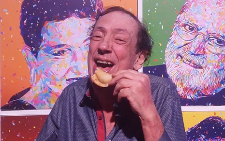 Marcos Oliveira sorri enquanto segura um pastel em frente a um mural com foto de comediantes