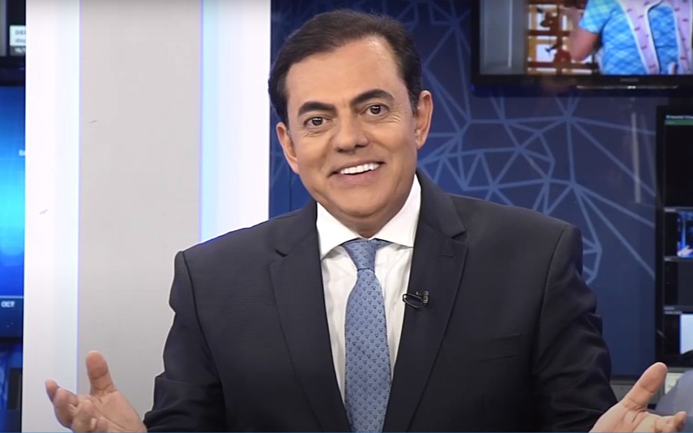 Marcos Tolentino sorrindo, vestindo terno e gravata, de braços abertos na bancada de um telejornal