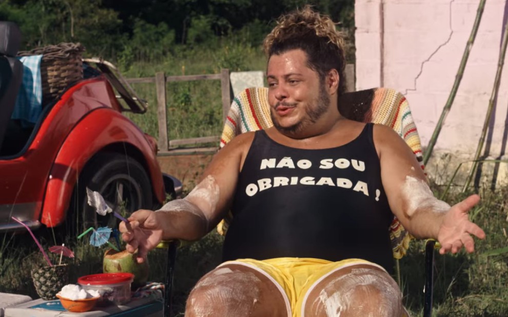 Marcus Majella sentado em cadeira de praia em cena de Vai que Cola 2 - O Começo
