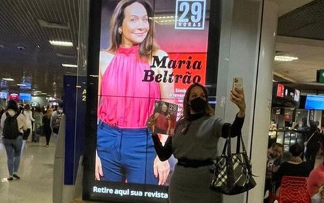 De máscara e com revista na mão em aeroporto, Maria Beltrão faz selfie com banner digital com sua imagem