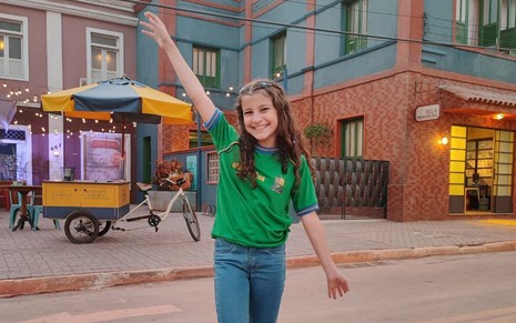 Caracterizada como sua personagem em Terra e Paixão, Maria Carolina Basilio posa com blusa verde, calça jeans e está com os braços abertos, sorridente