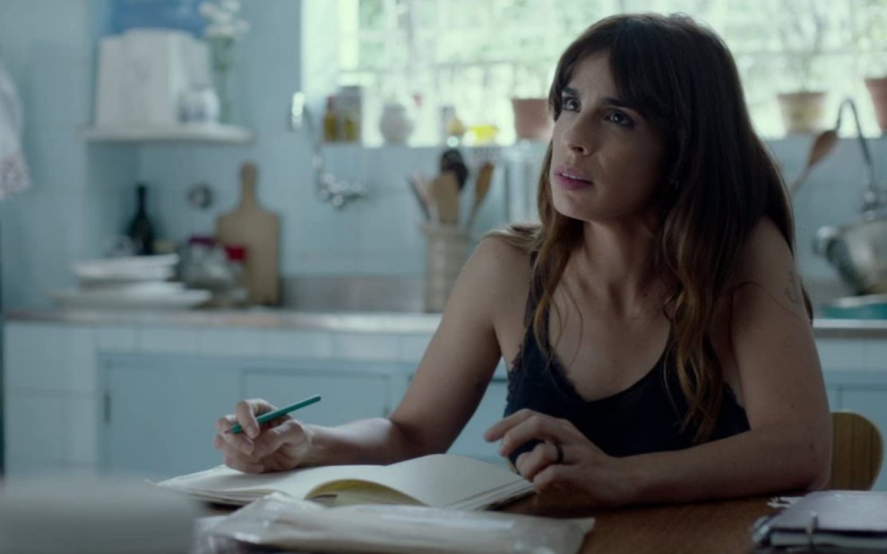 Maria Ribeiro sentada em uma mesa na cozinha, com um lápis na mão, o caderno sobre a mesa; ela apoia os braços e olha para algo