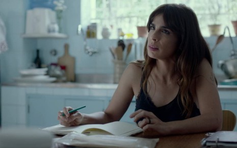 Maria Ribeiro sentada em uma mesa na cozinha, com um lápis na mão, o caderno sobre a mesa; ela apoia os braços e olha para algo