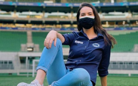 Mariana Spinelli sentada no gramado do estádio, calça jeans, camisa da Fox Sports e máscara preta