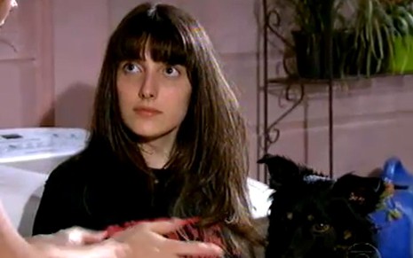 A atriz Clarice Falcão como Mariana em A Favorita; ela está sentada olhando para Catarina (Lilia Cabral), que não aparece na imagem, com o cachorro no colo