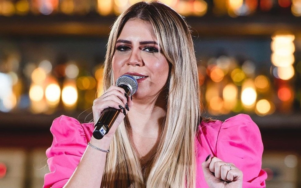 Marília Mendonça está com um microfone na mãe, vestida com uma camisa rosa, e canta em show