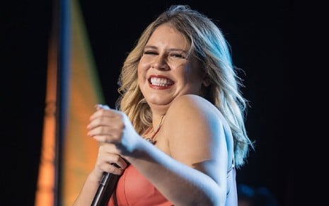 Marília Mendonça cantando em um show com uma camisa rosa e um microfone na mão