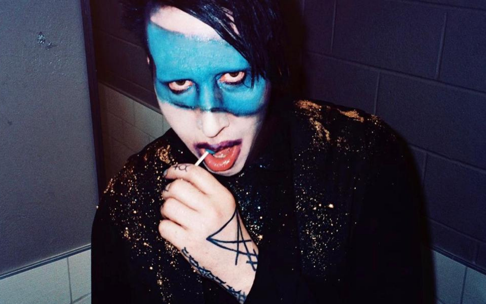 Marilyn Manson usa blusa de manga comprida preta com detalhes em dourado, chupa um pirulito e tem uma faixa azul pintada no rosto
