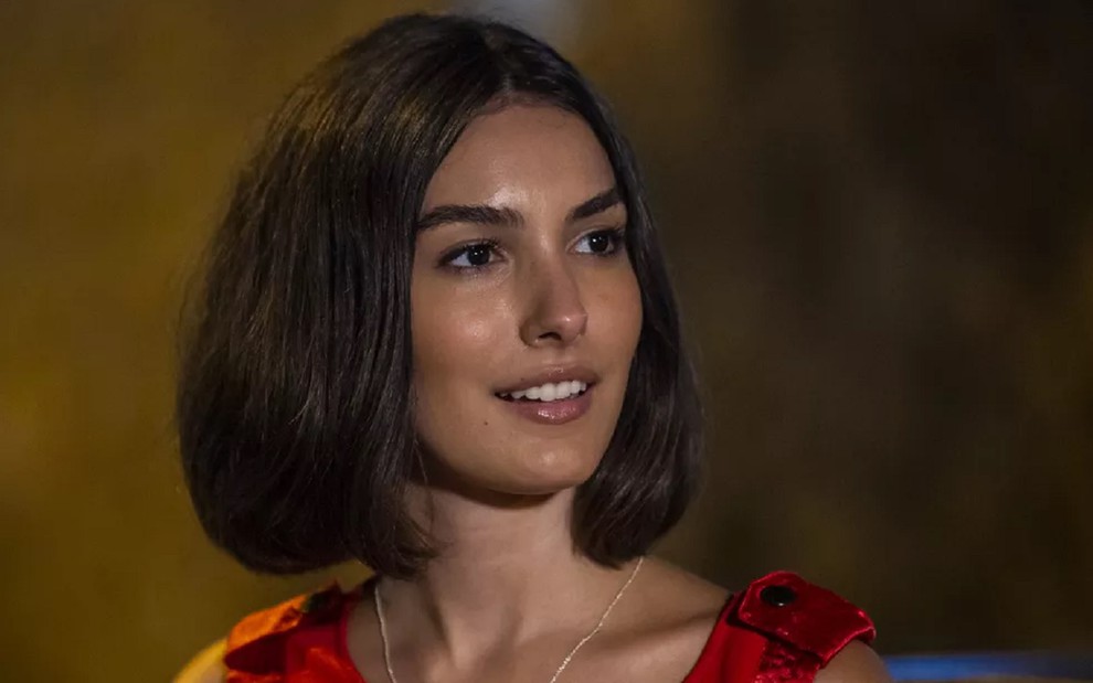 Marina Moschen usa um vestido vermelho e sorri em cena da novela Verão 90, seu último trabalho na Globo