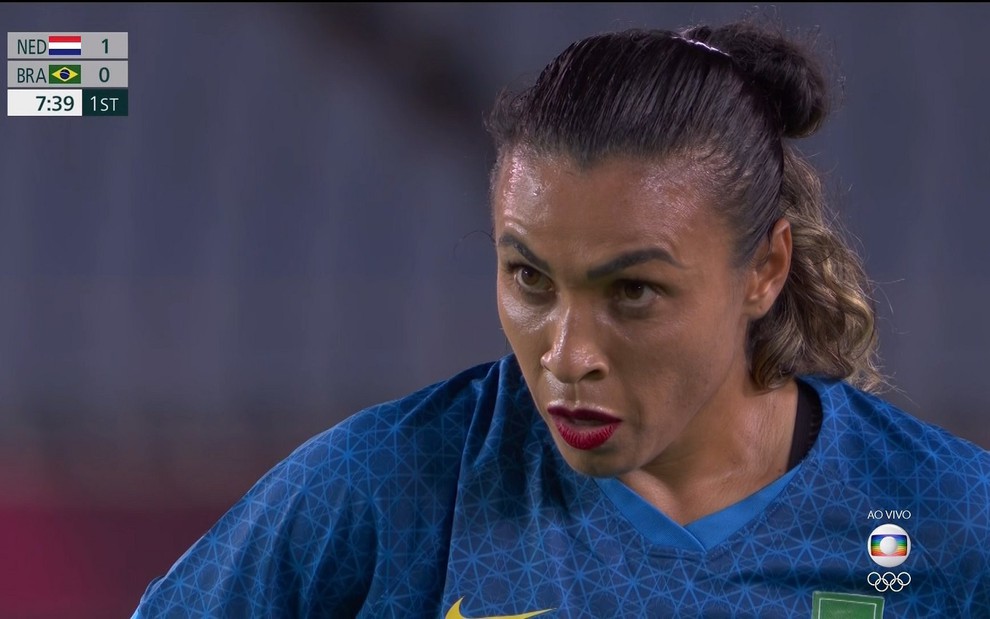 Marta olha focada para a goleira antes de bater pênalti. Ela usa uma camisa azul e um batom vermelho, além do cabelo preso