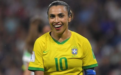 Marta sorri na Copa do Mundo. Ela usa uma camisa amarela e um batom vermelho, além do cabelo preso