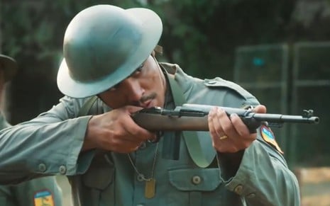 Matheus Dias está com roupa de soldado verde e aponta uma arma para frente em cena como Bento na novela Além da Ilusão