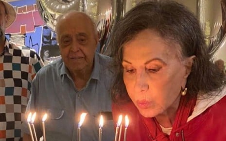 Os atores Mauro Mendonça e Rosamaria Murtinho durante a festa de aniversário de 90 anos da atriz; eles estão atrás do bolo enquanto ele a observa assoprar velinhas