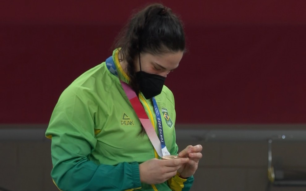 Mayra Aguiar com um casaco verde amarelo, de máscara preta e olhando para a sua medalha de bronze em Tóquio-2020
