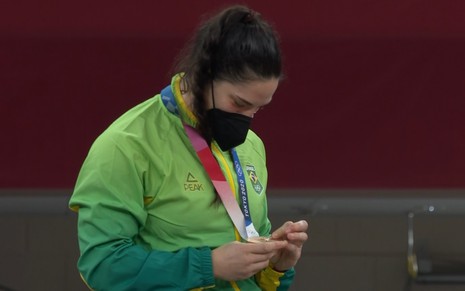 Mayra Aguiar com um casaco verde amarelo, de máscara preta e olhando para a sua medalha de bronze em Tóquio-2020