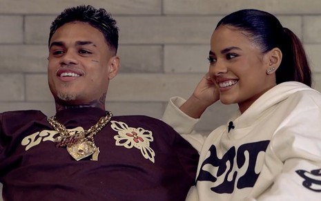 MC Cabelinho e Bella Campos estão sentados em um sofá e sorriem, em entrevista ao Fantástico, em maio