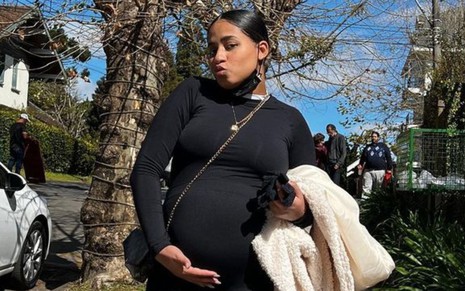 Imagem de MC Loma com a mão na barriga de grávida; ela usa roupa preta e cabelo preso