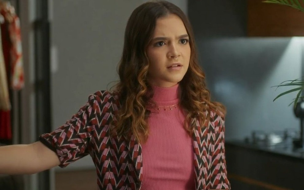Em cena de Vai na Fé, Mel Maia usa blusa rosa e camisa por cima estampada; ela está falando com alguém com expressão de surpresa