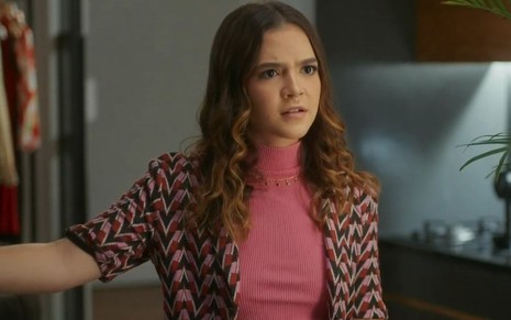 Em cena de Vai na Fé, Mel Maia usa blusa rosa e camisa por cima estampada; ela está falando com alguém com expressão de surpresa