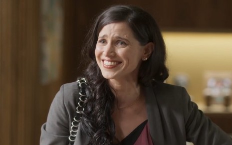 Mel Lisboa grava cena como Regina em Cara e Coragem; ela sorri e olha para o lado em cenário da novela