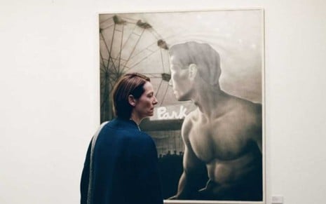 A atriz Tilda Swinton está de costas, e seu rosto aparece apenas de perfil, enquanto olha para a fotografia de um homem com o torso nu em frente a uma roda gigante em cena de Memória
