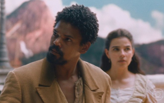 Jorge/Samuel (Michel Gomes) olha para o lado com expressão de revolta; Pilar (Gabriela Medvedovski) aparece ao fundo da cena de Nos Tempos do Imperador