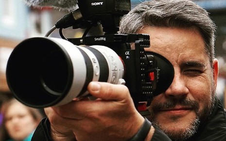 Mikael Fox, repórter acuasado de assédio na Globo, usa uma câmera e foca nela em uma filmagem