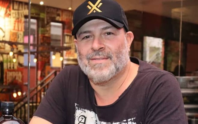 Mingau usa uma blusa e um boné pretos; ele dá um leve sorriso para a câmera. A barba e as sobrancelhas estão grisalhas.