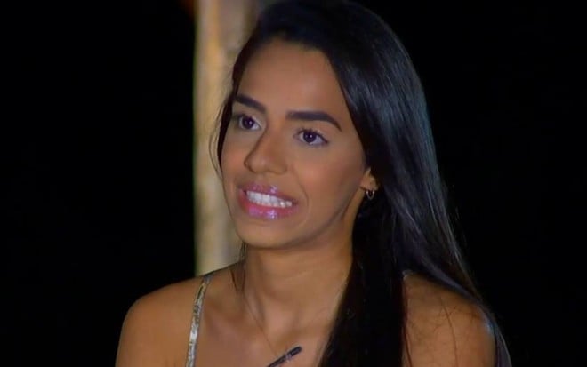 A câmera está focada no rosto de Mirella, ela está sorrindo de forma forçada, está com o cabelo solto e veste regata na cor clara