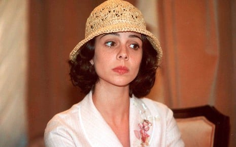 Miriam Freeland com expressão desconfiada, chapéu de palha e vestido branco, em cena de O Cravo e a Rosa