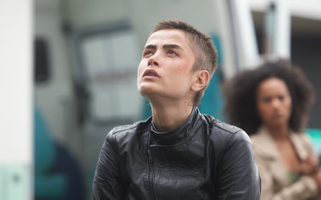 Maria Casadevall está com a cabeça raspada e jaqueta de couro em cena de A Garota da Moto. Ela olha para cima, com expressão pensativa