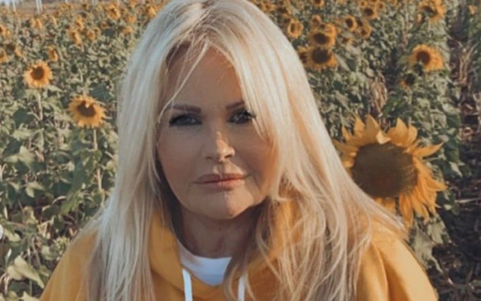 A ex-modelo Monique Evans com moletom laranja, expressão séria, posa para foto em ambiente externo, com sol e plantas ao fundo
