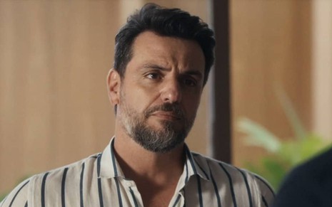 Em cena de Travessia, Rodrigo Lombardi está usando uma blusa azul com listras finas brancas e está com a expressão de descontentamento