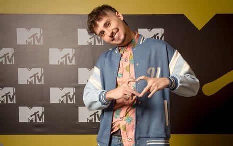 Leo David posa diante de um backdrop da MTV e faz um coraçãozinho com as mãos com o pescoço todo torto