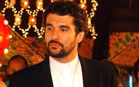 Nicola Siri caracterizado como padre Pedro em Mulheres Apaixonadas (2003), de terno e camisa branca, olha para a esquerda