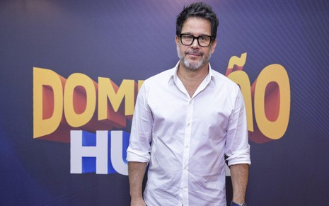 Murilo Benício está com blusa branca em frente ao painel com a marca Domingão com Huck