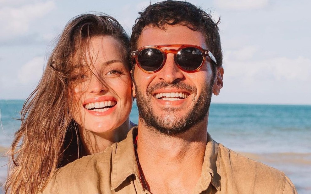 Flavia Lucini sorri, com o rosto colado ao de Leandro Lima; ele, que está na frente da noiva, também está sorridente e usa um óculos escuros. Ao fundo, é possível ver uma praia.