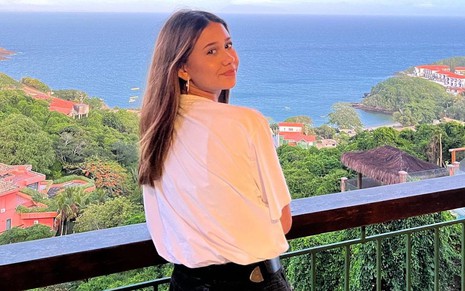 Nathália Costa posa com blusa branca em uma varanda que dá para ver o mar no fundo